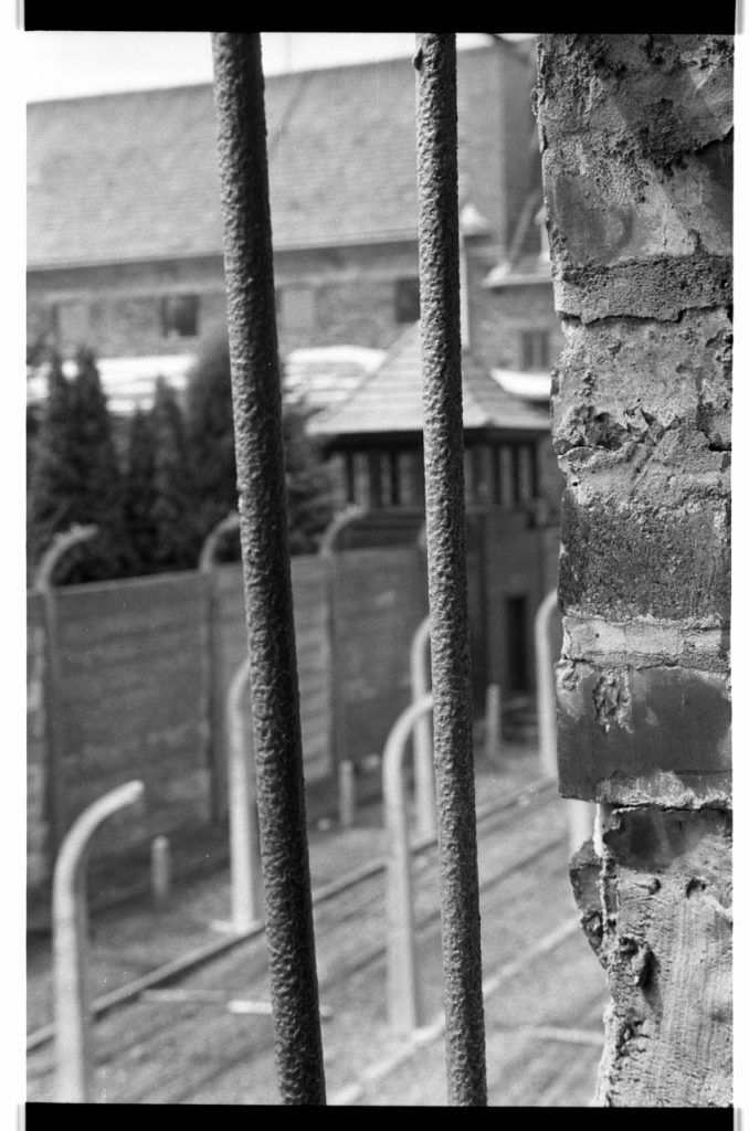 B/W photo of rusty bars. In the background are fence posts and a gatehouse. Nikon D70 Film Camera 2004 - Negative Scan from Nikon COOLSCAN V ED 2008