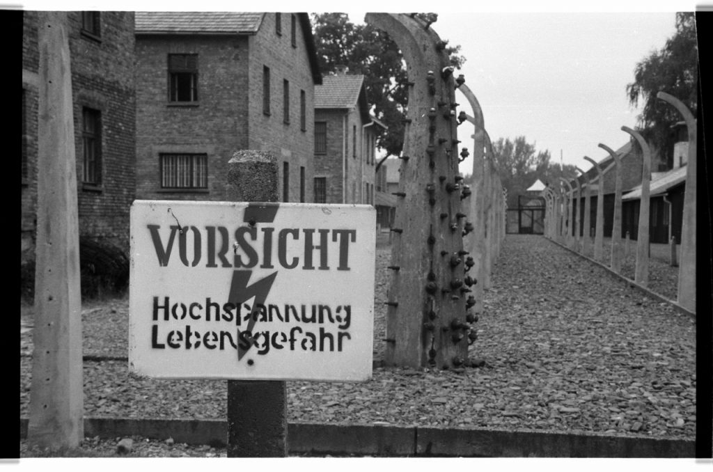 B/W photo of a Vorsicht warning sign. In the background is fence posts and brick buildings.  Nikon D70 Film Camera 2004 - Negative Scan from Nikon COOLSCAN V ED 2008