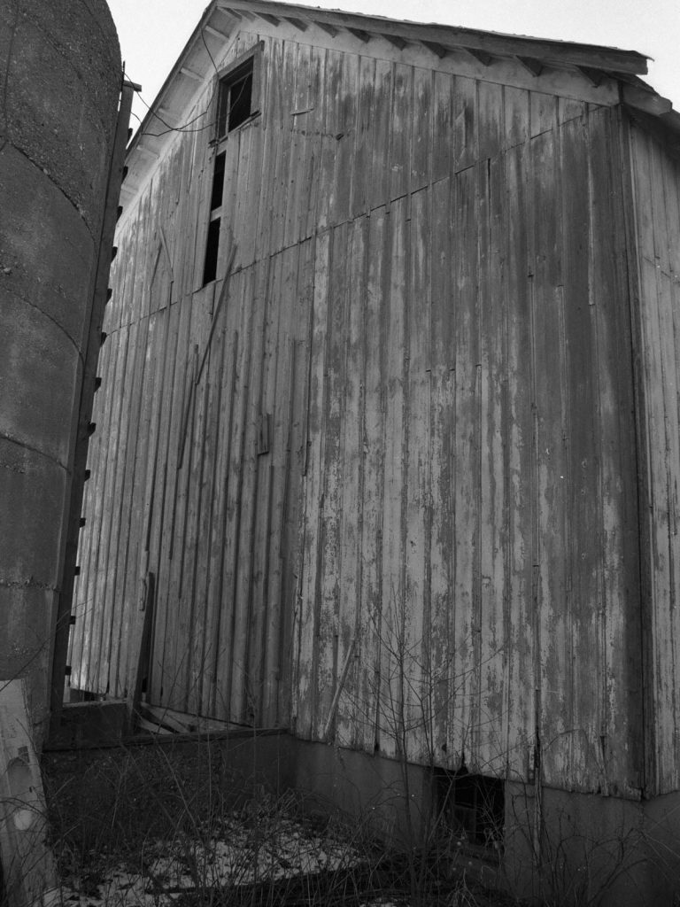 B/W photograph of a white run-down barn and a concrete silo. There is snow on the ground.